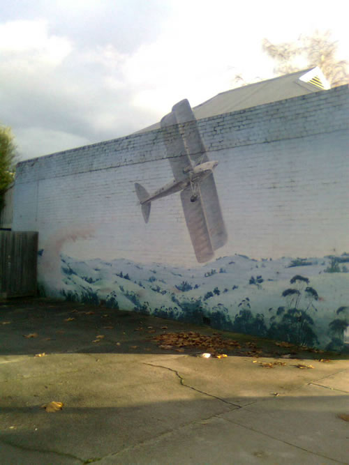 Mural on the side of a shop with a small biplane in flight. The artist has extended the wing up above the wall line using wood or some other material.