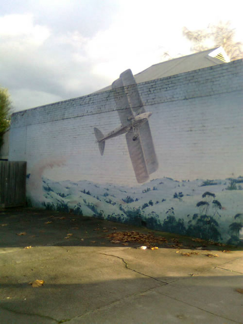 MirbooMurals_Plane2 Mural on the side of a shop with a small biplane in flight. The artist has extended the wing up above the wall line using wood or some other material.