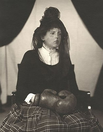 Woman sitting in a parlour or sitting room, wearing nineteenth century clothes, black veil and boxing gloves