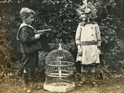 Little boy aiming a large toy (we hope) gun at a little girl, who is smiling happily. There is a bird cage on the ground between them.