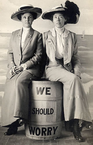 Two women in late C19th or early C20th dress sitting on a barrel with the words "We should Worry"painted on it